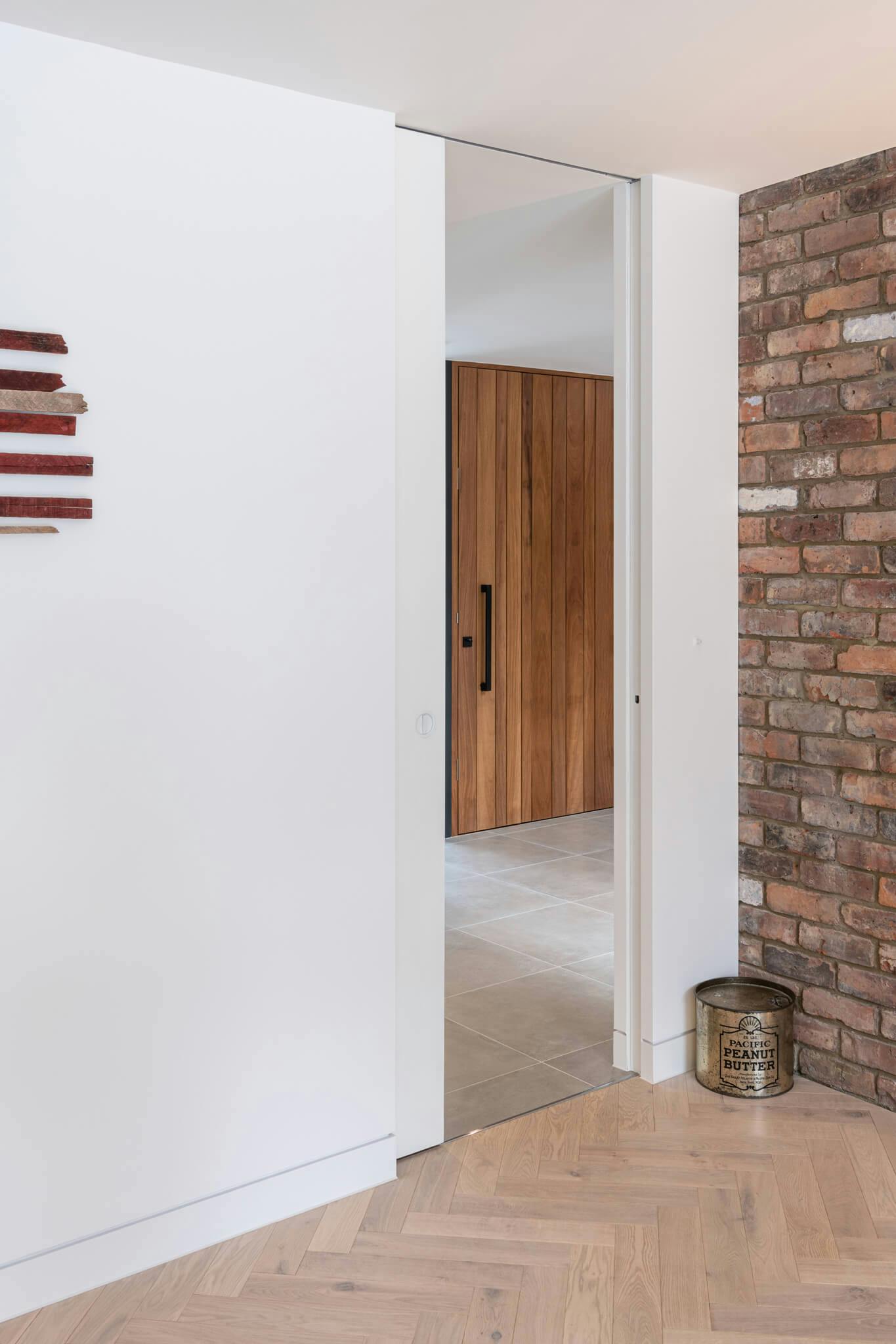 a contemporary white walled interior with wooden floor and brick wall with a white sliding pocket door that leads into a reception with tiled floor.