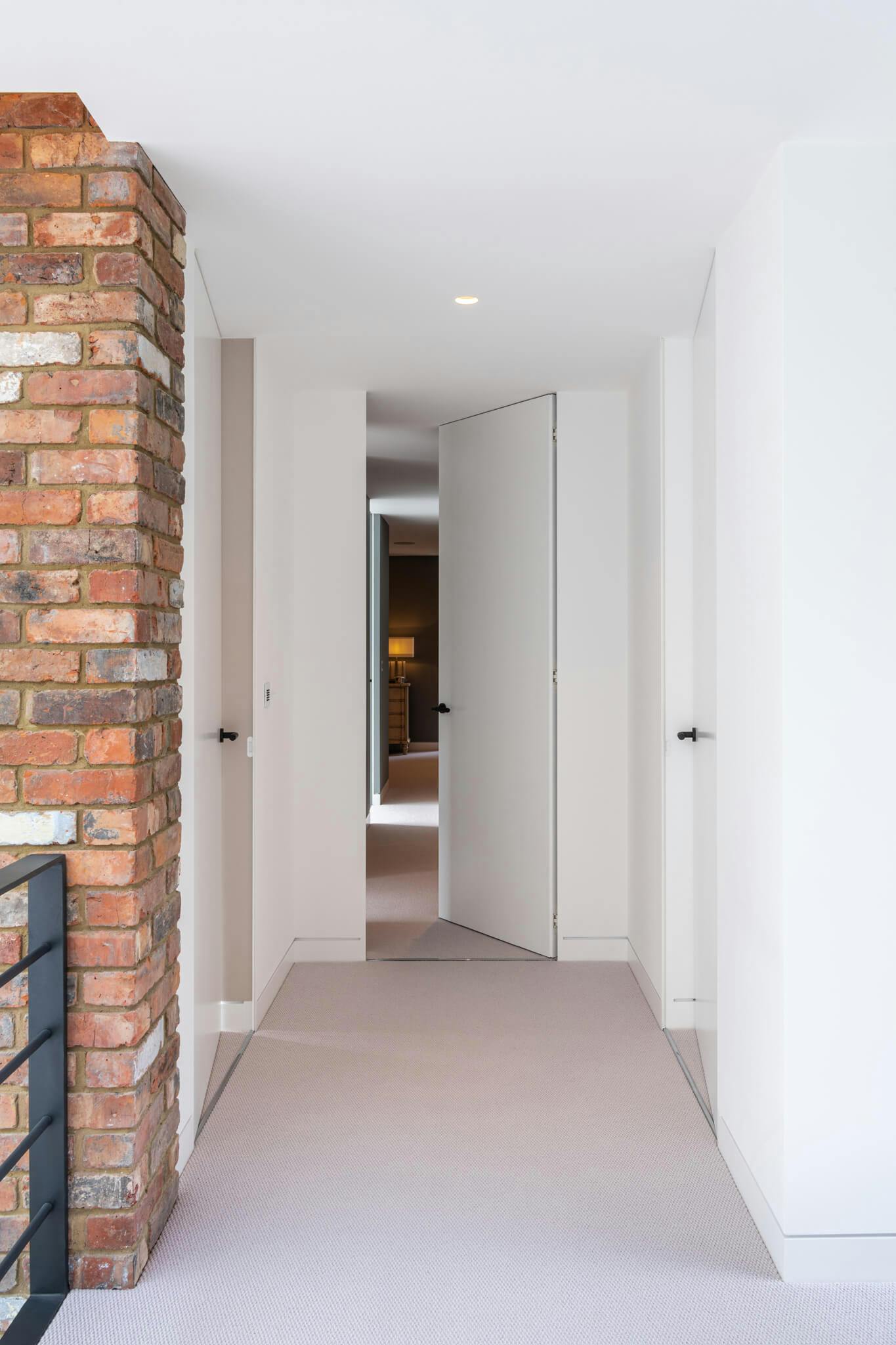 A carpeted corridor with a flush door at the end leading into a bedroom.  Foreground with steel rail, brick feature wall and white walls and doors.