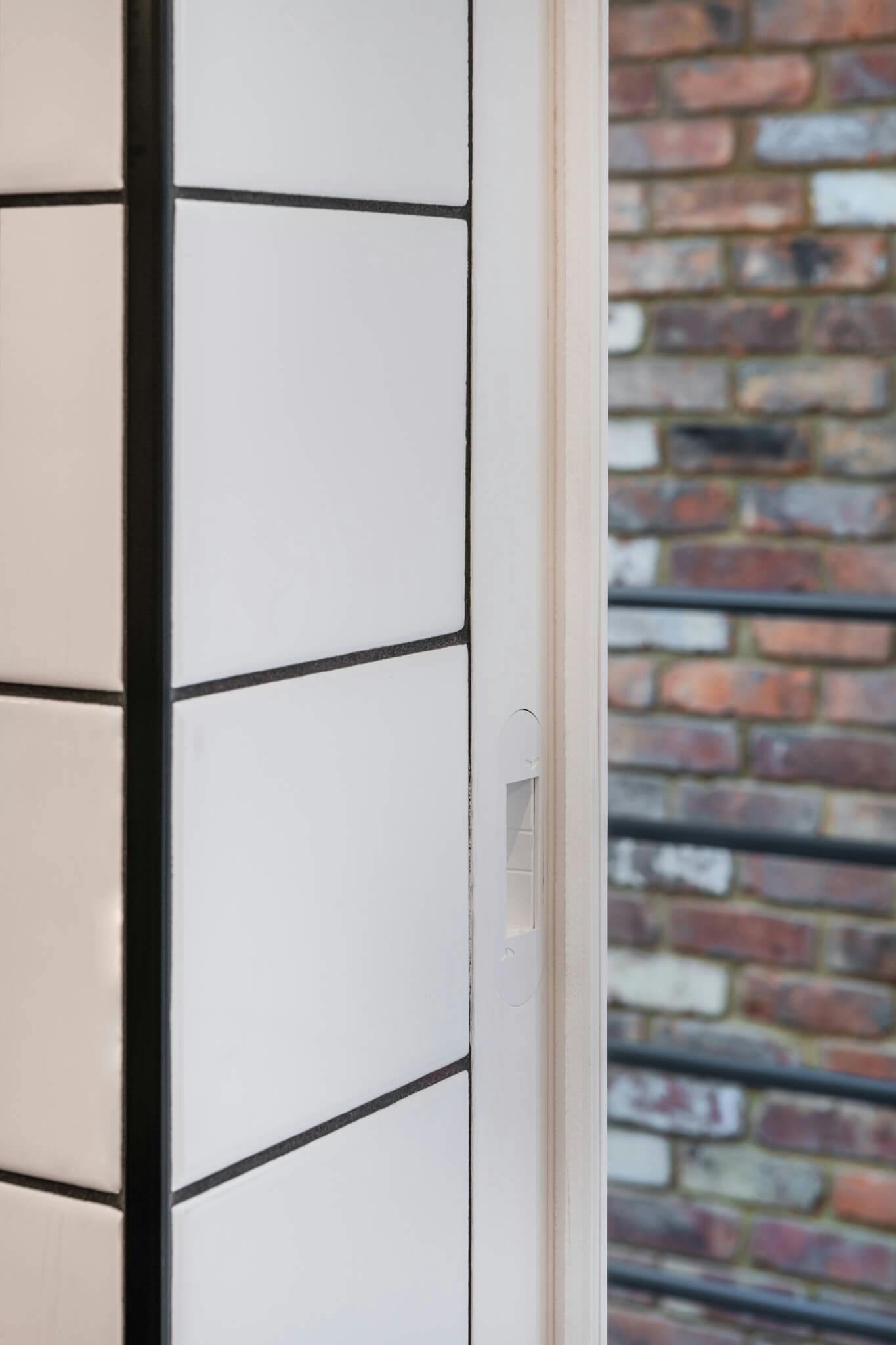 A white flush door post with white keep set in white tiled opening with black grout with a brick wall in background.