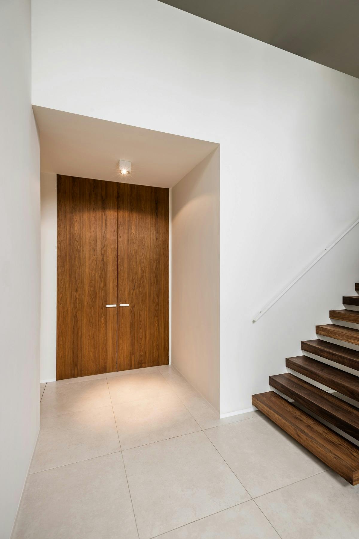 A tiled hallway with brown floating stairs leading out of the frame to the right, white walls and a set of closed double flush brown wooden doors with silver handles.
