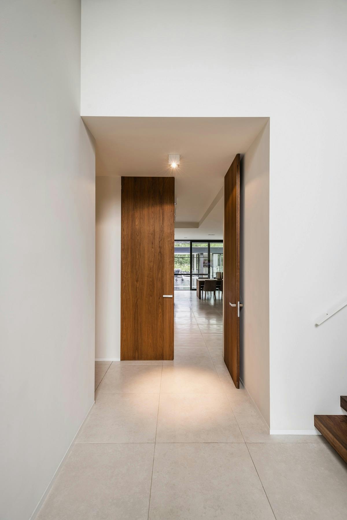 A white reception with tiled floor and foot of wooden stairs, with corridor leading to wooden double flush doors with one door open leading to living area.