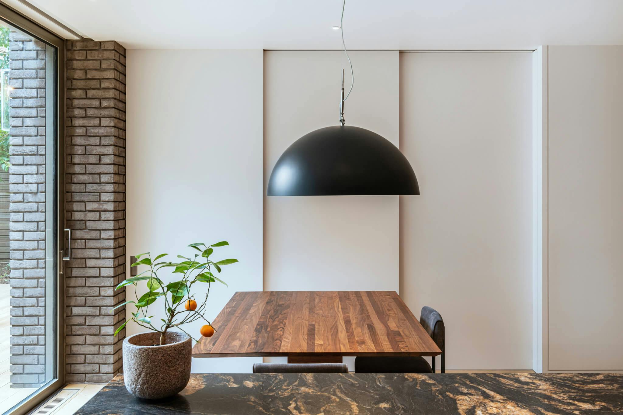 A view from the kitchen of a large metal dome hanging light with an orange plant, wooden table and a triple set of telescopic sliding doors.