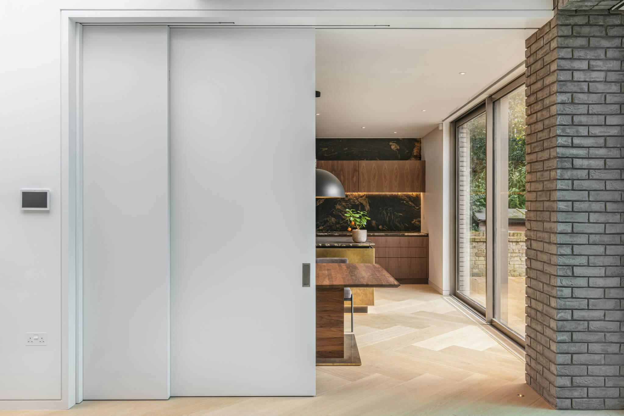 A set of half open white sliding telescopic doors leading into a wood and marble kitchen with wooden flooring and a dark grey brick wall to the right.