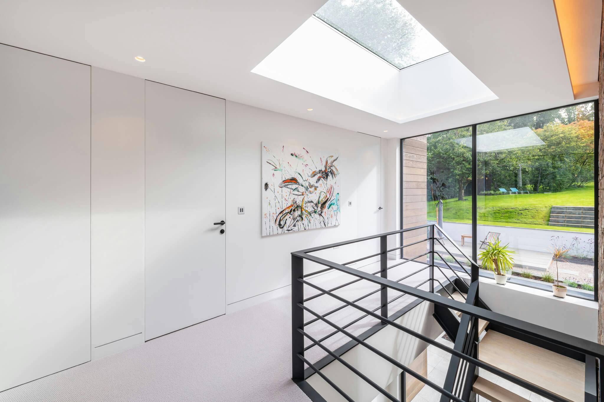 A large neutral coloured landing area with a view to the garden through steel glazed window panes and two large closed white flush doors with an abstract canvas painting.