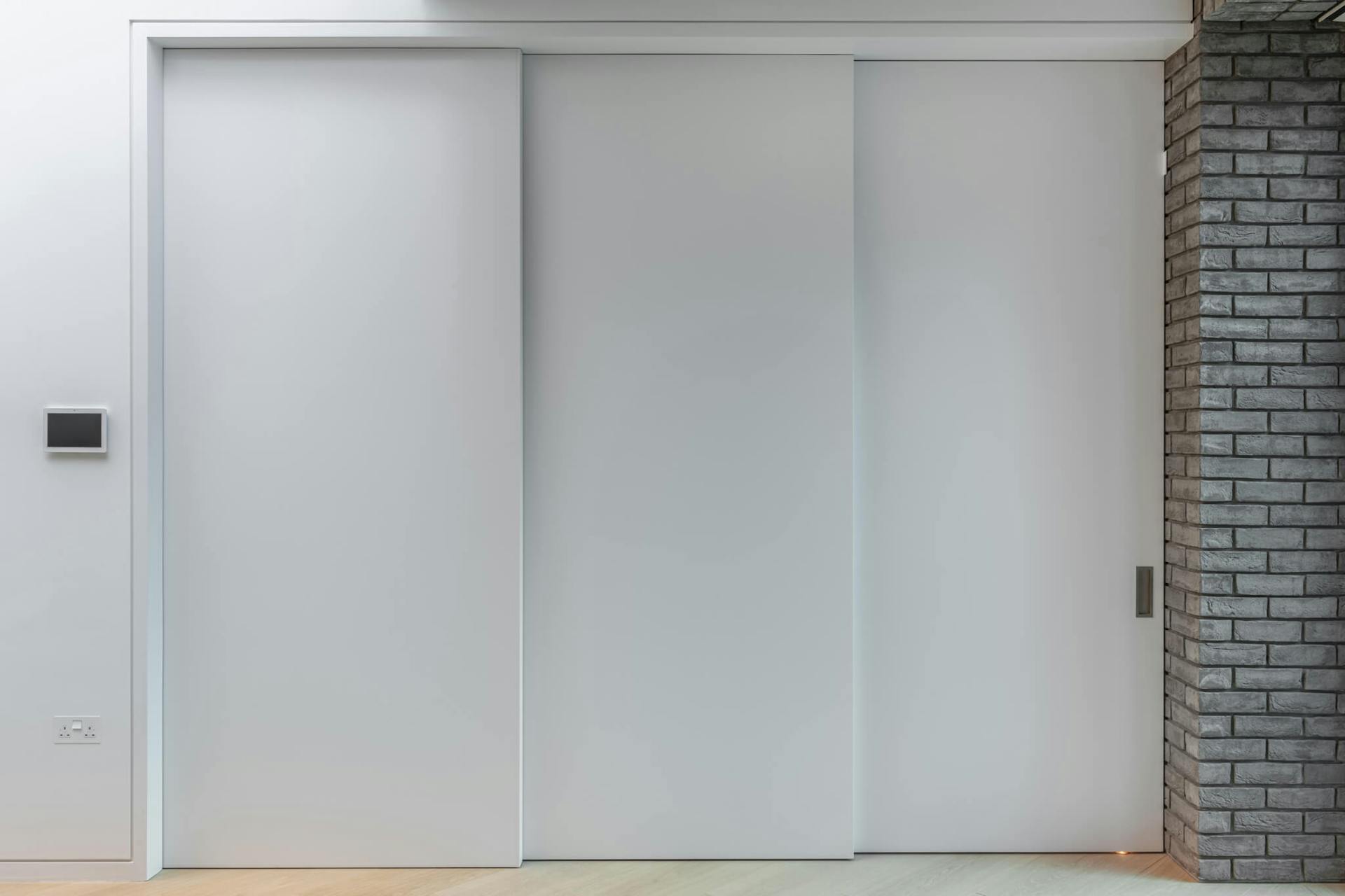 A fully closed set of three white sliding doors on a telescopic track against a grey brick internal wall.