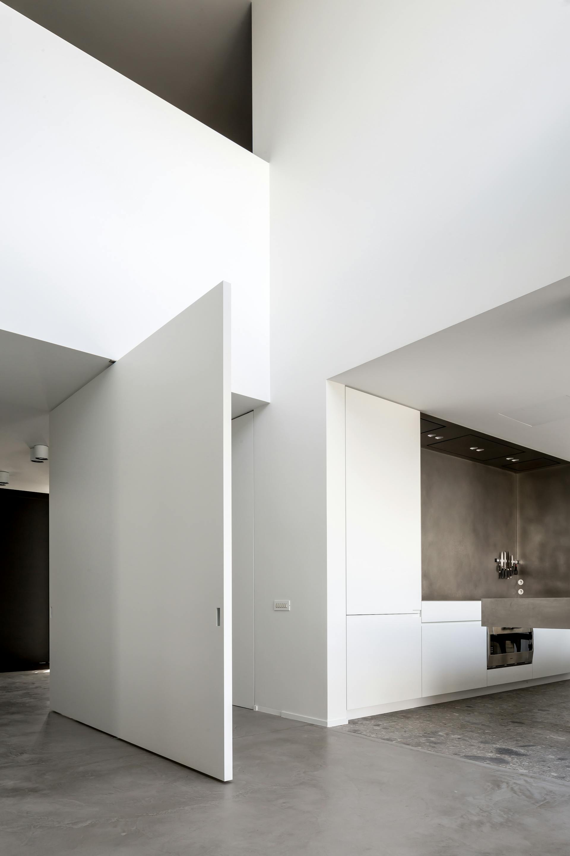A huge white centrally pivoting door fully open in a large white space with concrete floor and kitchen worktops.