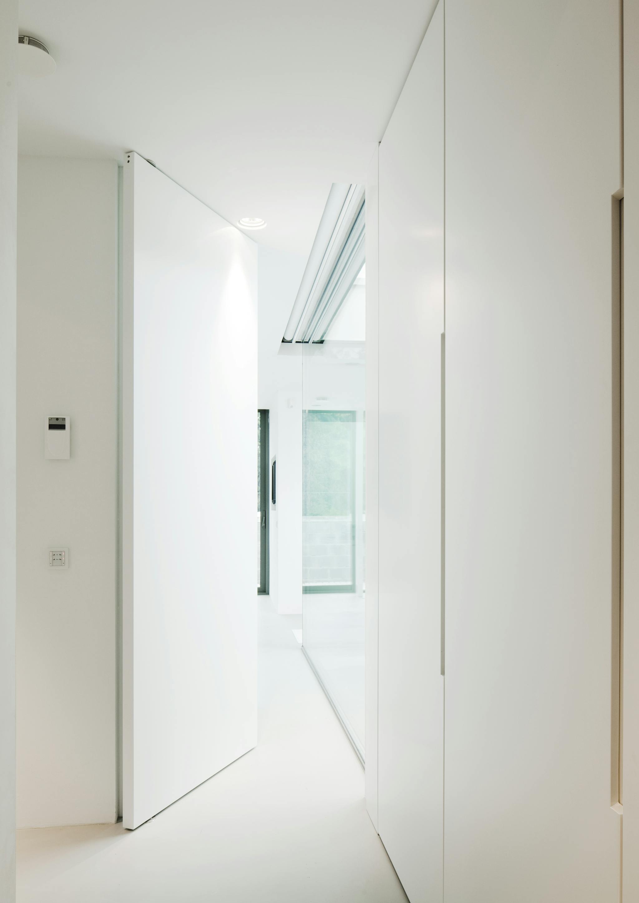 A large pivoting flush door slightly ajar leading into a corridor with full height glazing on the right hand side.
