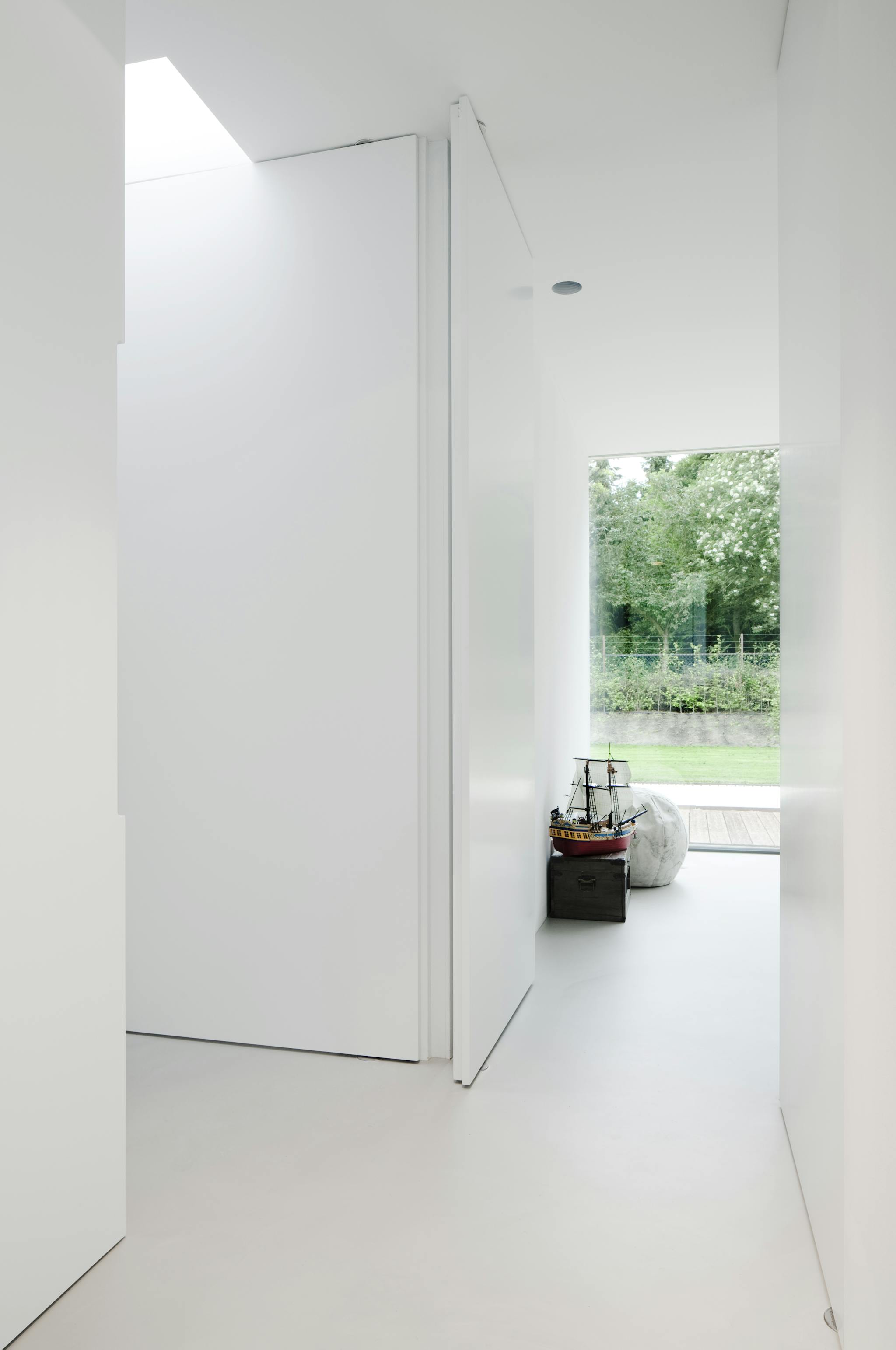 A white minimal interior with closed pivoting door and opening into a bedroom.
