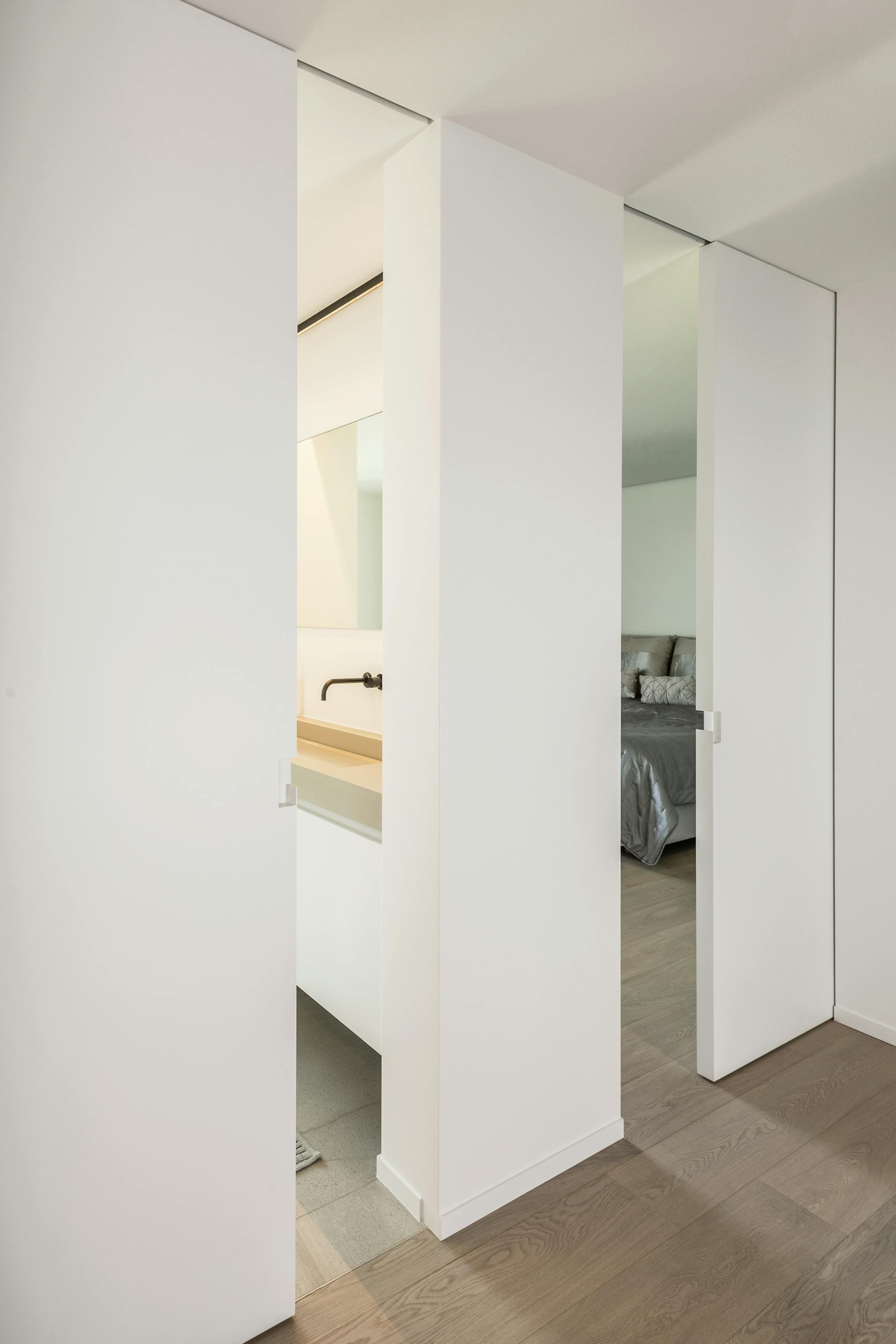 Two white single sliding pocket doors in a private home leading to a bathroom and bedroom, with dark wooden flooring and white minimal interior
