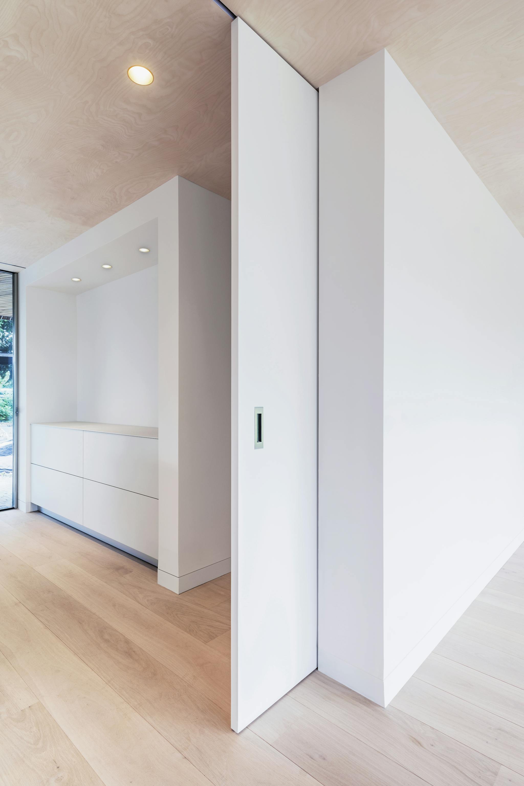 A minimalist interior with light wood flooring and a large white sliding pocket door with a recessed pull