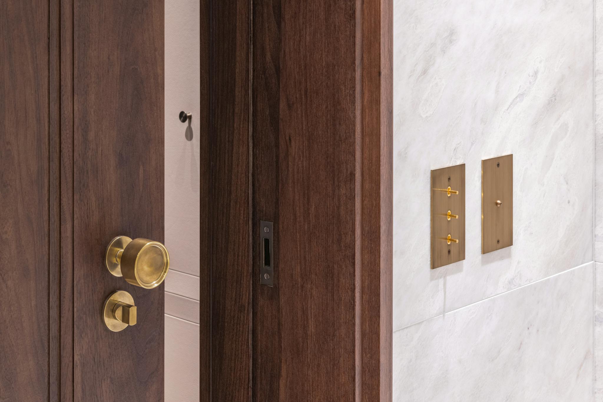 Dark wood door frame with heavy set door with solid brass ironmongery against a pale tiled wall and electrical toggle plates.
